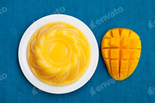 Preview: Mango pudding, jelly, dessert on white plate. Blue textile background. Top view.