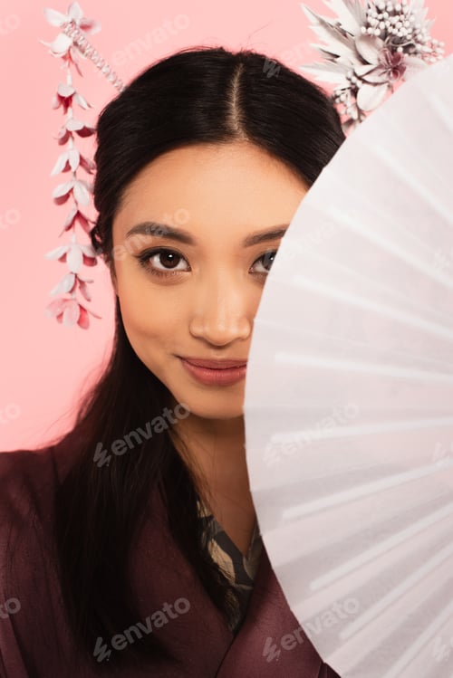 Preview: Young asian woman looking at camera while holding fan isolated on pink