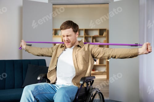 Preview: Young disabled man in wheelchair doing exercises with rubber band at home.