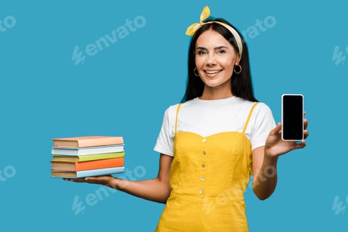 Preview: positive woman holding books and smartphone with blank screen isolated on blue