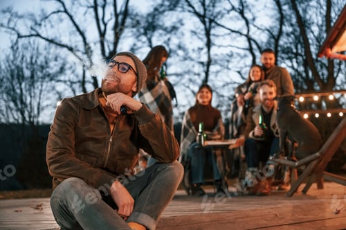Preview: Man is smoking. Group of people is spending time together on the backyard at evening time
