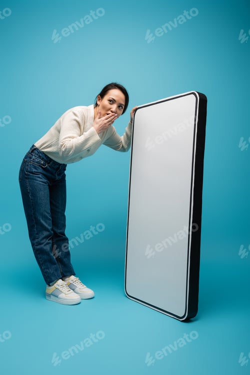 Preview: Shocked asian woman covering mouth near big smartphone model on blue background