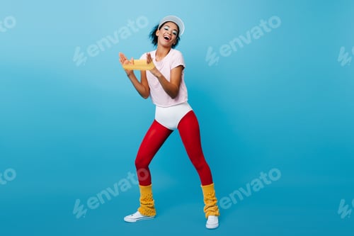 Preview: Laughing black woman training with expander. African fitness girl using sport equipment on blue bac