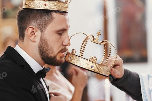 Preview: Bride and groom kissing golden crowns from priest