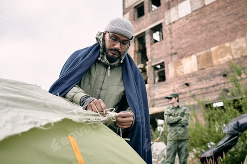Preview: Male refugee pitching tent against half ruined building