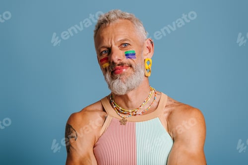 Preview: Portrait of mature gay man with rainbow flag make-up looking at camera against blue background