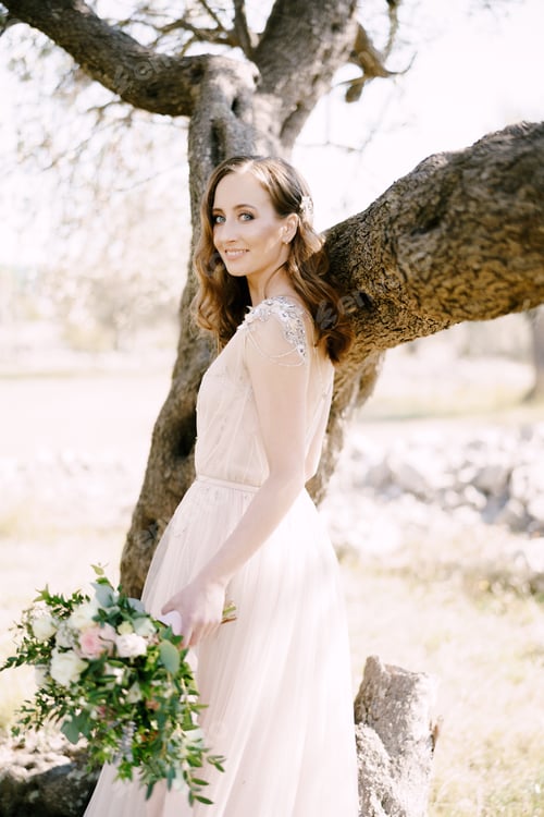 Preview: Bride with a bouquet stands half-turned near a tree trunk
