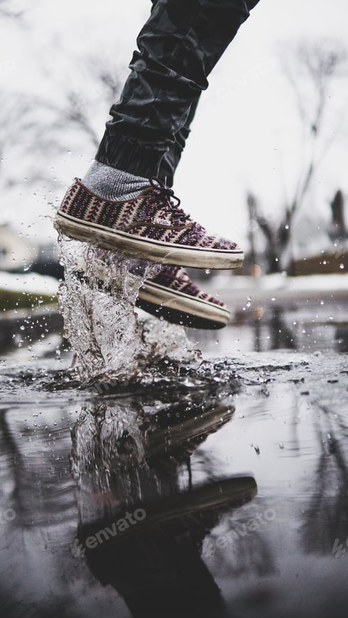 Preview: Vertical shot of person in black jeans and low-top sneakers jumping on puddle and splashing all over