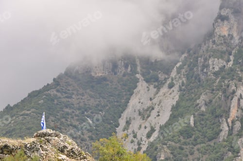 Preview: Greek flag in distance amongst misty mountains
