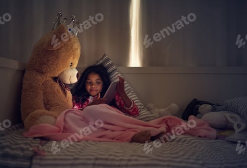 Preview: Shot of a little girl using a digital tablet while lying in bed with her teddy at night
