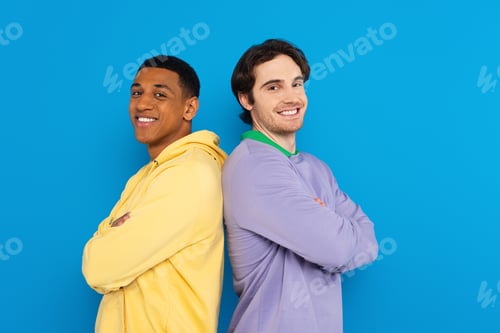 Preview: smiling interracial friends in back to back pose isolated on blue