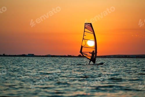 Preview: Still sea surface, man practicing wind surfing and golden sunset in sky