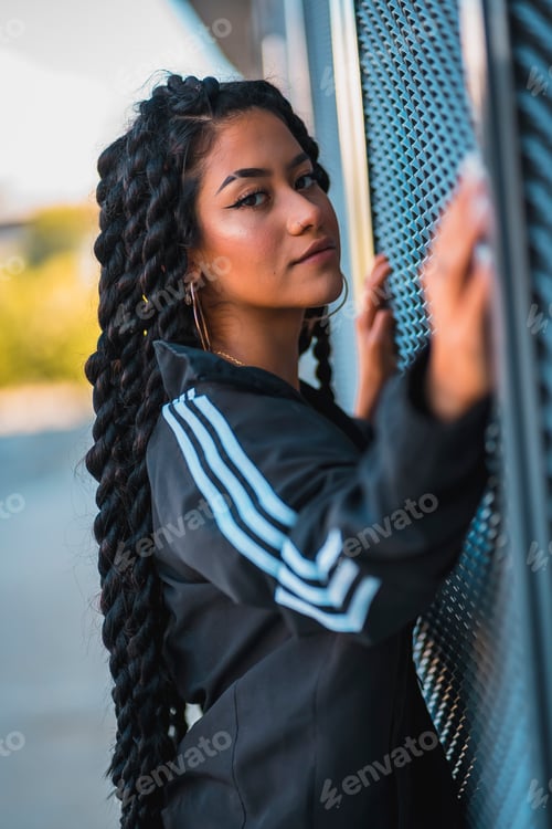 Preview: Urban session. Young black ethnic woman with long braids and with tattoos, looking glass, smiling