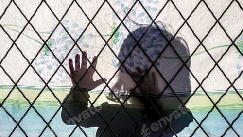 Preview: Shadow of a little girl on a colorful fabric behind a chain-link fence under sunlight