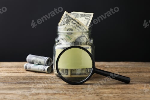 Preview: Jar with blank note, money and magnifying glass on wooden table against black background