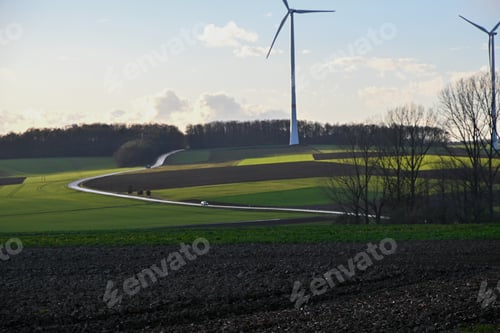 Preview: Narrow road with cars by a wind power plant against a blue cloudy sky on a sunny day