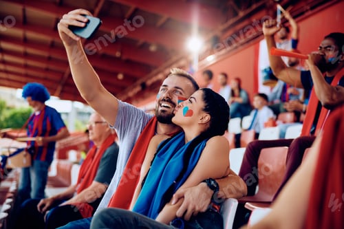 Preview: Young couple having fun while taking selfie during sports game at the stadium.