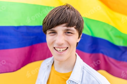 Preview: Young gay man smiling at camera over rainbow flag background.