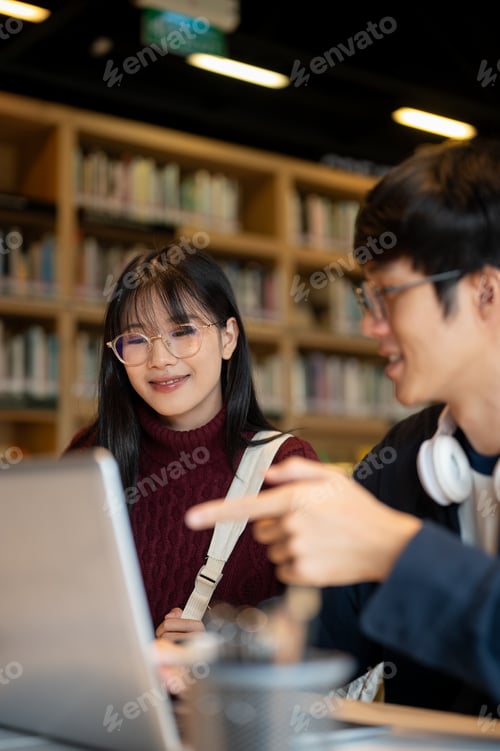 Preview: Glasses asian woman student looking at laptop of a man friend while sitting at table in library.