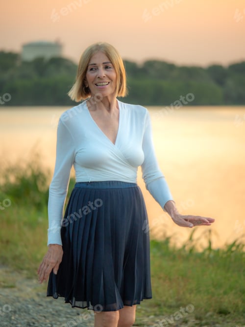 Preview: caucasian senior woman wearing white shirt and pleated skirt in a riverbank at dusk