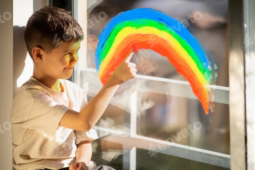 Preview: Cheerful small european boy draws rainbow with finger on window, enjoy spare time in children room