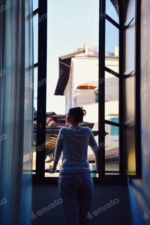 Preview: Woman is looking out a large open window overlooking the rooftops of a city in Europe