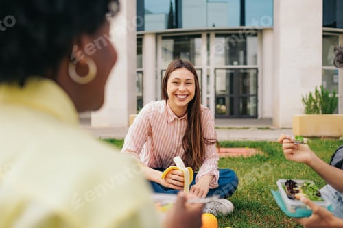 Preview: Beautiful latin student eating fruit with friends sitting on the grass of college campus.