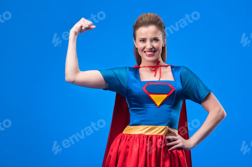Preview: portrait of smiling woman in superhero costume showing muscles isolated on blue