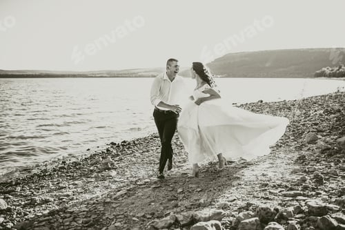 Preview: bride and groom laughing run together against rays of setting sun in summer