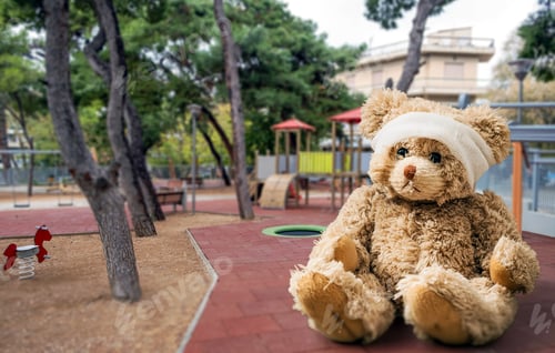 Preview: Teddy bear with bandage on head sitting alone on grass at childrens playground.