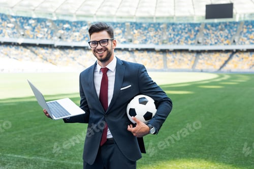 Preview: smiling young businessman in suit with laptop and soccer ball sitting on football pitch at stadium,