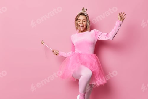 Preview: Cheerful young woman dances carefree has upbeat mood wears small crown and dress holds magic wand en