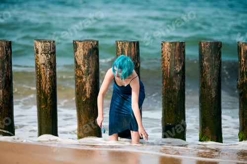 Preview: Beautiful blue-haired woman performance artist in blue dress standing on beach holding paint brush