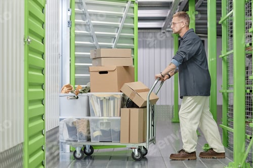 Preview: Smiling Female Client Loading Boxes into Self Storage Unit Using Cart at Warehouse