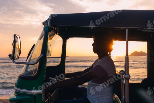 Preview: Silhouette of tuk tuk driver driving along coastal road during ocean sunset in Sri Lanka