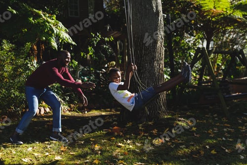 Preview: African american father with son having fun and playing on swing in garden