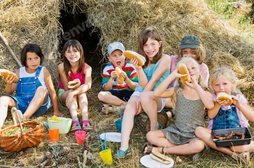 Preview: A group of children enjoying a picnic with hot dogs in front of a hay bale on a sunny day.