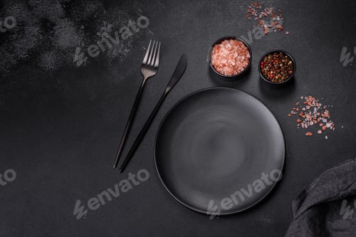 Preview: An empty black plate on a dark concrete background with spices, herbs and cutlery