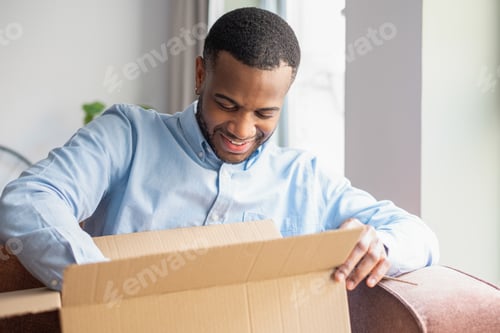 Preview: Portrait of a smiling African American man sits on sofa at home unpacking a parcel