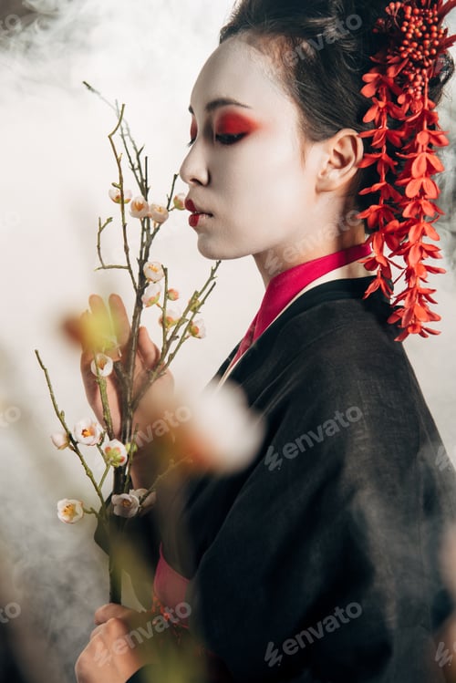 Preview: selective focus of beautiful geisha in black kimono with red flowers in hair and sakura branches in