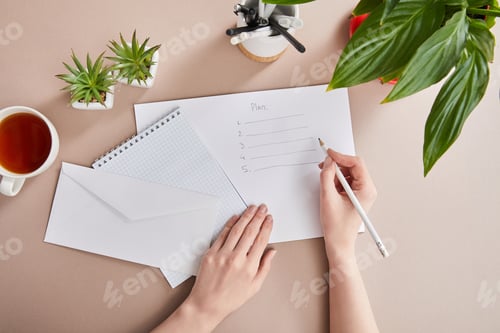 Preview: cropped view of woman writing plan on paper near green plants, cup of tea, envelope, blank notebook