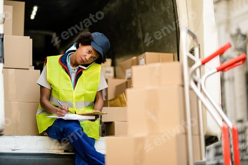 Preview: Black female courier writing notes while communicating on mobile phone in a van.