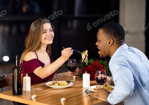 Preview: Cheerful interracial couple having fun at dinner in restaurant, woman feeding boyfriend