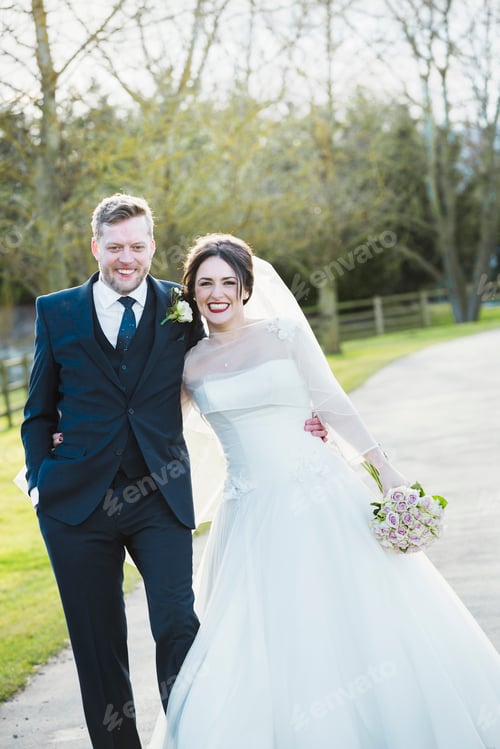Preview: A bride and groom on their wedding day walking arm in arm down a path in the sunshine laughing.