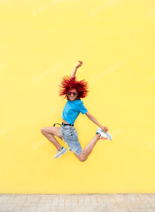 Preview: vertical photo of a happy latin woman with red afro hair jumping over isolated yellow wall