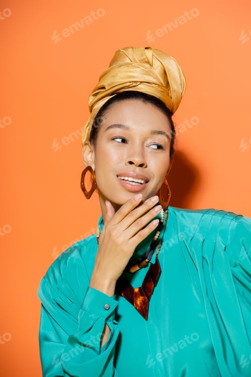 Preview: Smiling african american woman in golden headscarf touching chin on orange background