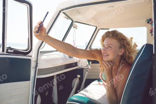 Preview: Beautiful Caucasian woman taking selfie with mobile phone while lying on seat in camper van at beach