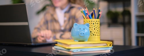Preview: blue piggy bank and textbooks on the table, in the background a teenager girl does homework on a