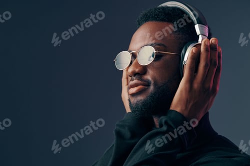 Preview: Young Black man enjoying music with headphones on, wearing stylish sunglasses, set against a dark