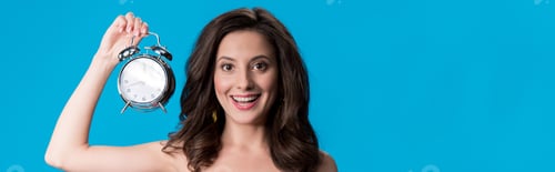 Preview: panoramic shot of happy elegant young woman in dress holding silver alarm clock isolated on blue